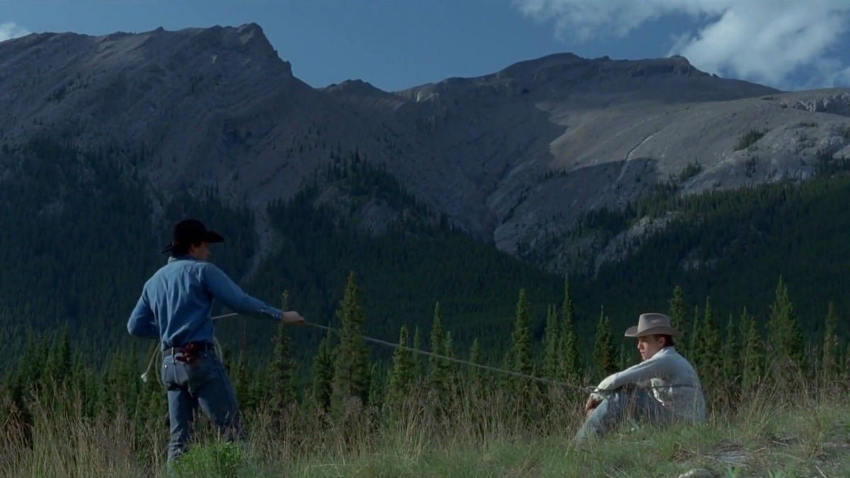 Two cowboys pass the time with a lasso beneath forested mountains.