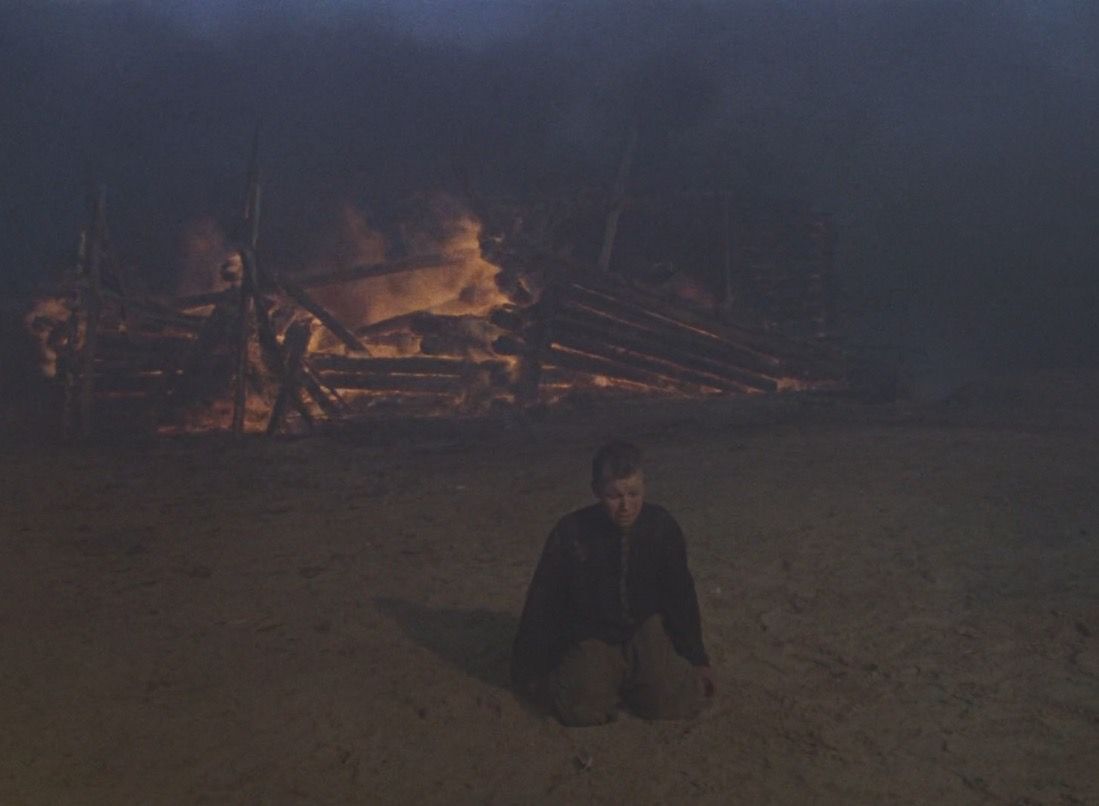 A boy kneels in front of a burning village.