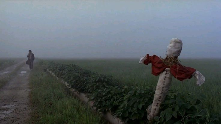 A scarecrow stands beside a misty field and a rural path.