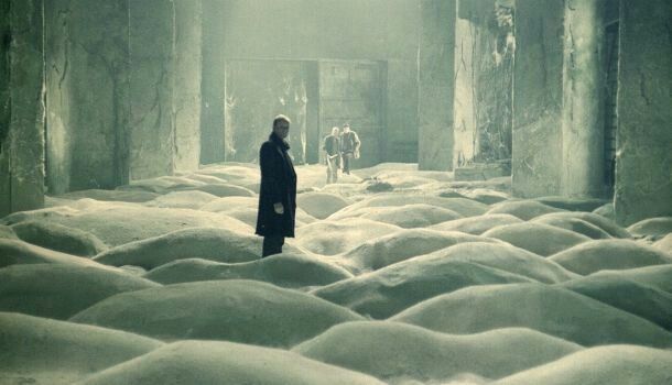 Travelers stand among sand dunes inside a ruined industrial room.