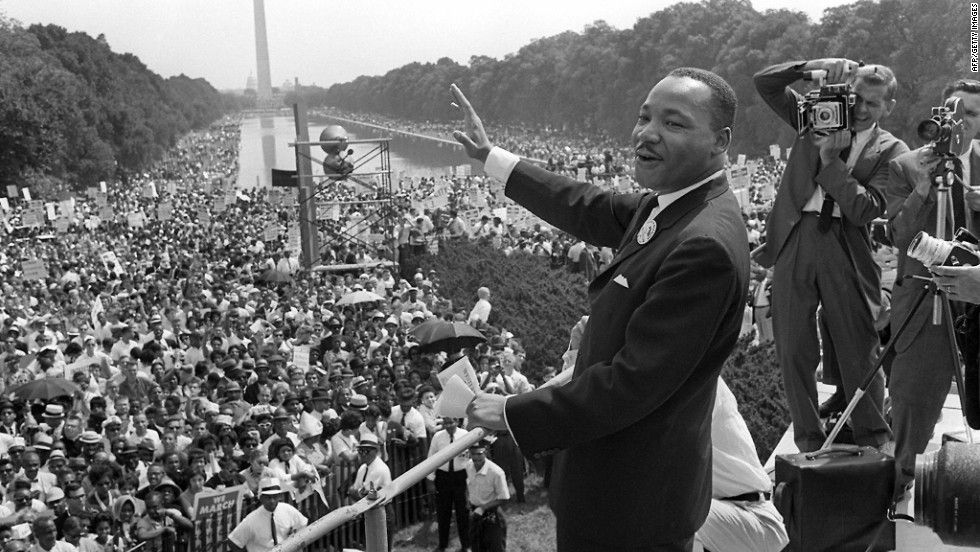 Black-and-white photograph of a speaker facing a large crowd beside the Reflecting Pool, with the Washington Monument in the distance.